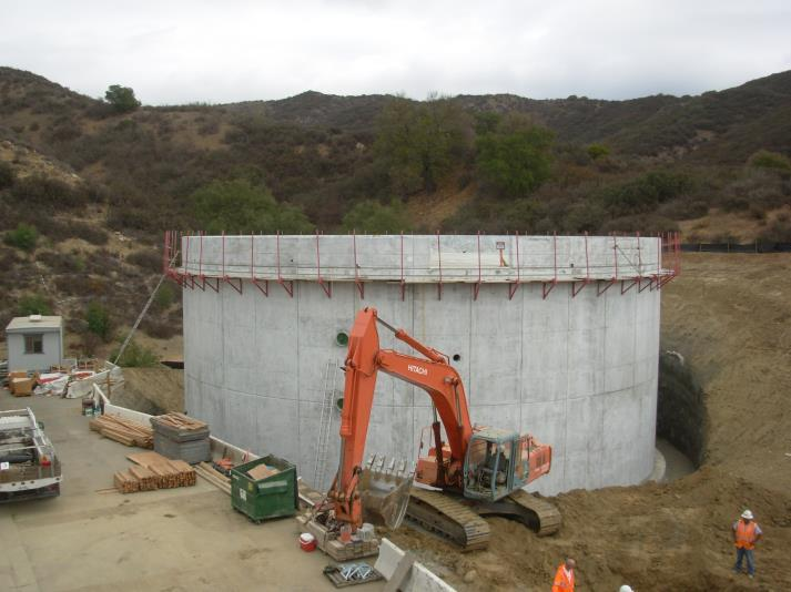 Construction site with a large concrete structure, an excavator, two workers, and materials in a hilly area.