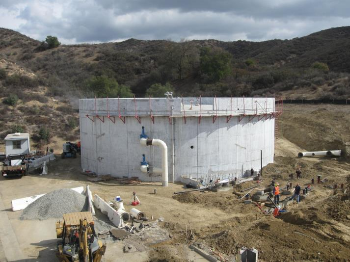A large cylindrical concrete structure under construction with workers and vehicles in a hilly area.