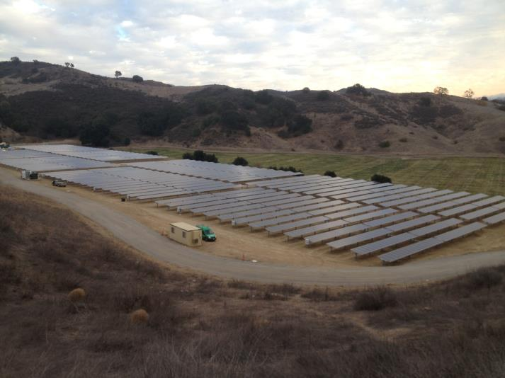 A solar farm with rows of solar panels in a hilly, grassy landscape under a partly cloudy sky.