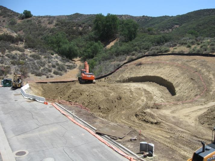 Construction site with an excavator working on a hilly terrain, surrounded by trees and dirt mounds.