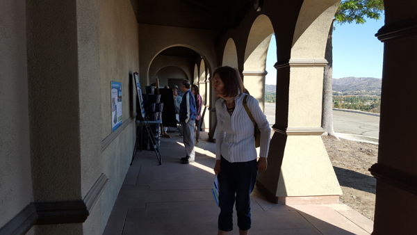 People walk under an arched walkway, examining display boards on a wall, with hills visible in the background.