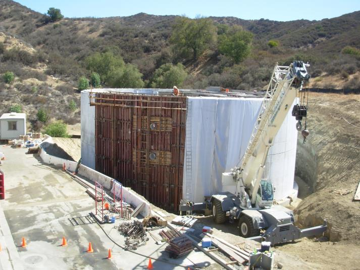 Construction site with a large concrete wall, a crane, and scattered building materials in a hilly landscape.