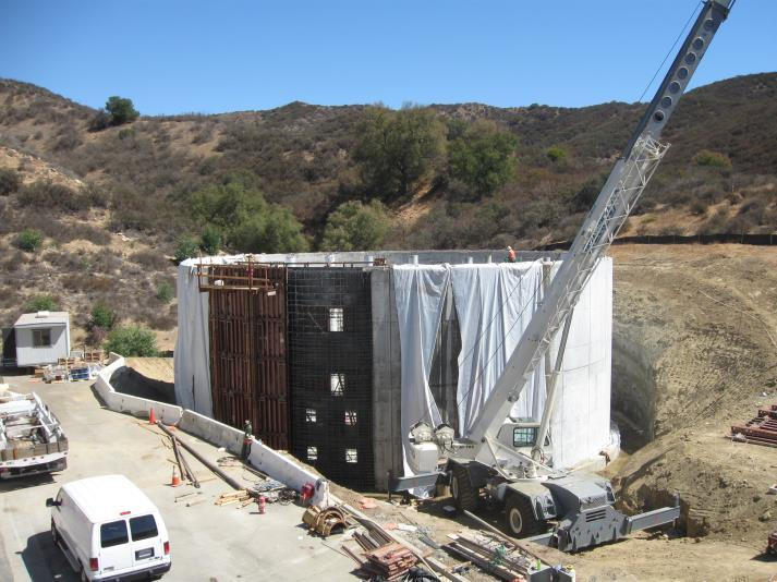 A construction site with a crane, partially built structure covered with tarps, and surrounding hills.