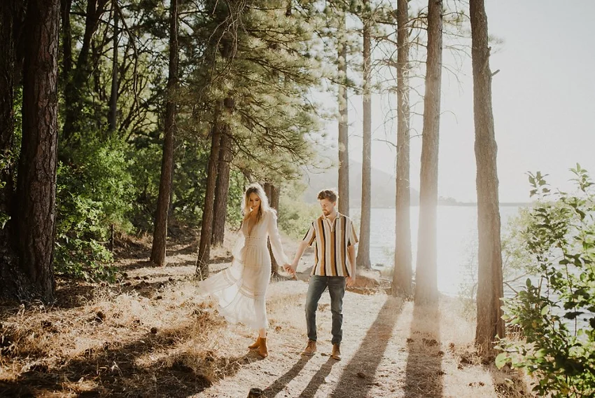 A couple holding hands, walking through a sunlit forest near a lake.