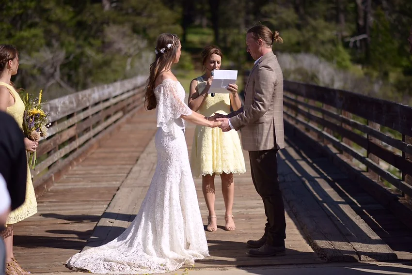 A wedding ceremony on a wooden bridge, with a bride and groom holding hands, and an officiant reading from a paper.
