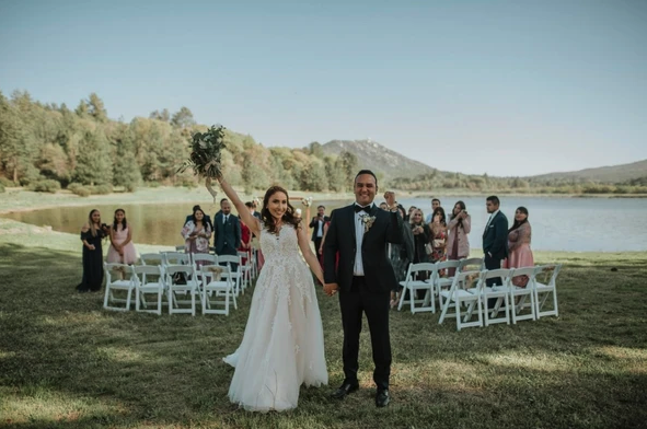 A couple celebrates in front of wedding guests outdoors by a lake, surrounded by trees and mountains.