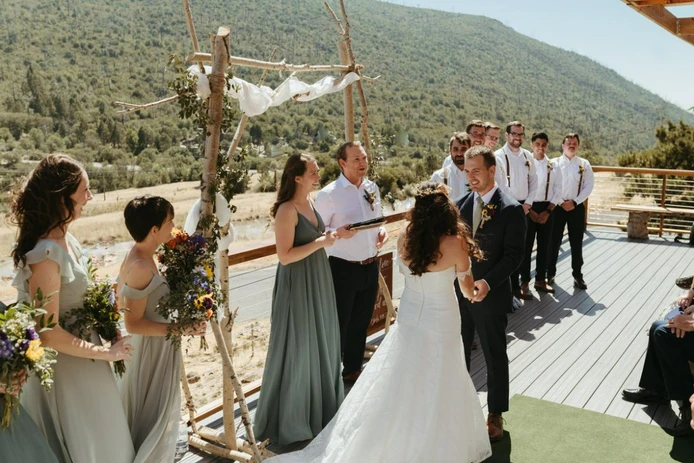 An outdoor wedding ceremony with a bride, groom, and bridal party on a wooden deck with mountain scenery.