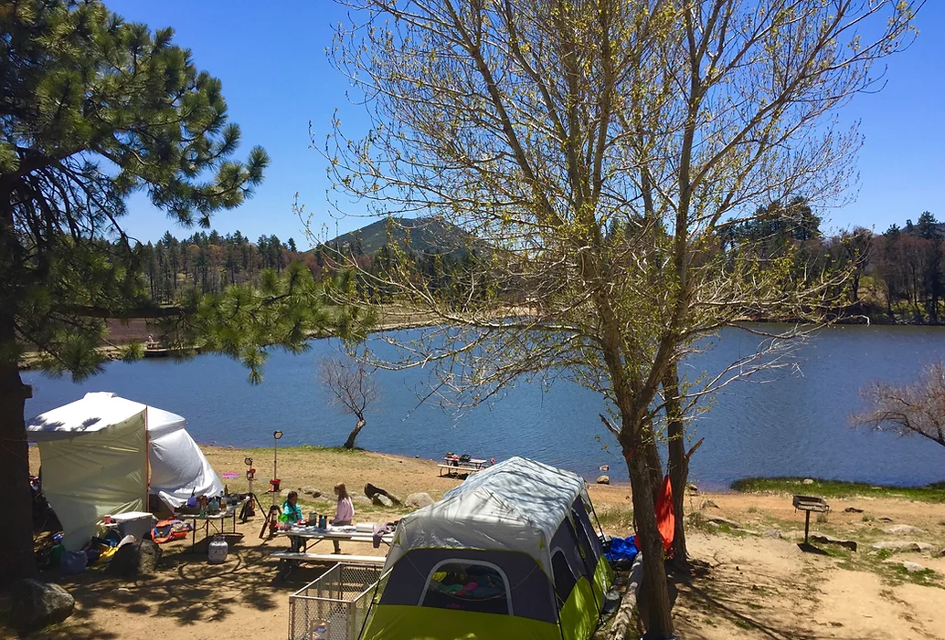 A scenic lakeside campsite with tents, picnic tables, and people under trees, surrounded by nature and a blue sky.