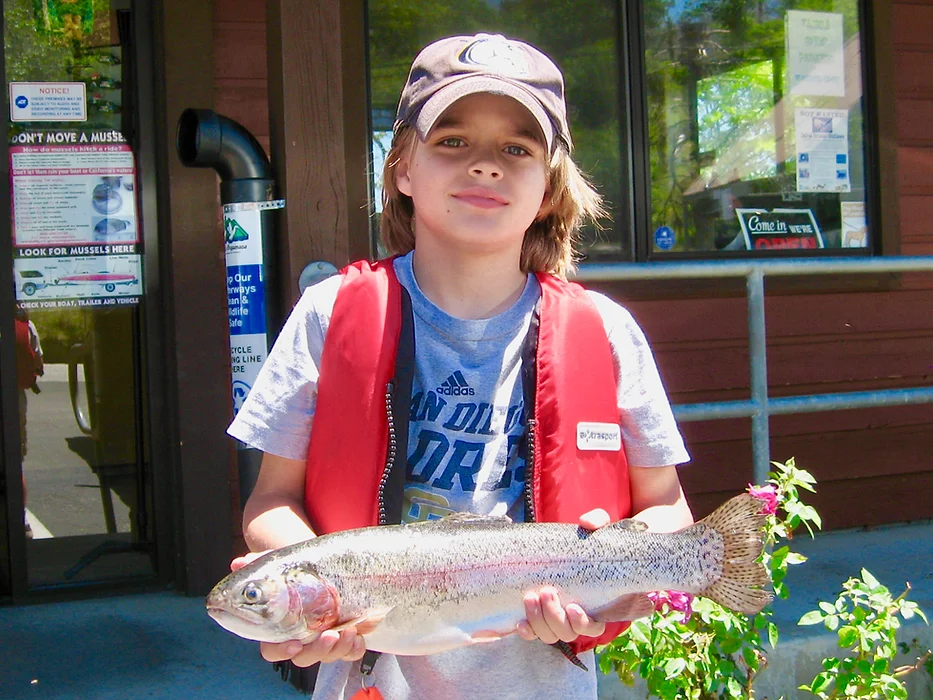 A kid in a red life jacket holds a large fish, standing outside a building on a sunny day.