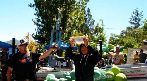 Two people celebrate at an outdoor event, holding a trophy and a fish amidst a crowd and festive decorations.