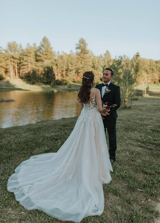 A couple standing by a pond, with the woman in a wedding dress holding flowers.