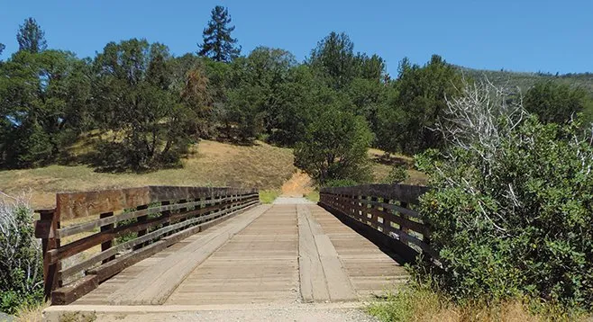 A wooden bridge spans a path, surrounded by trees and greenery under a clear blue sky.