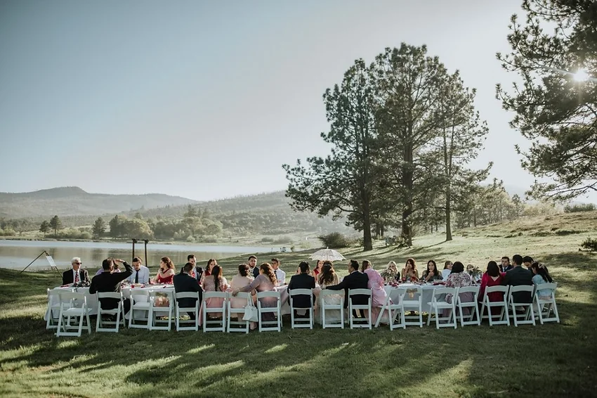 Outdoor gathering with people seated around a long table, set in a scenic landscape with trees and a lake.