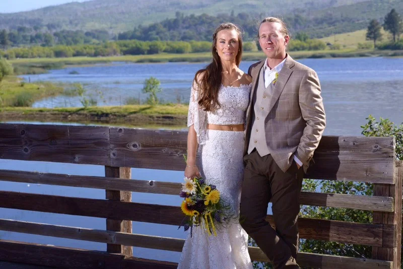 A couple in wedding attire poses by a wooden fence, with a lake and green hills in the background. One person holds a bouquet.