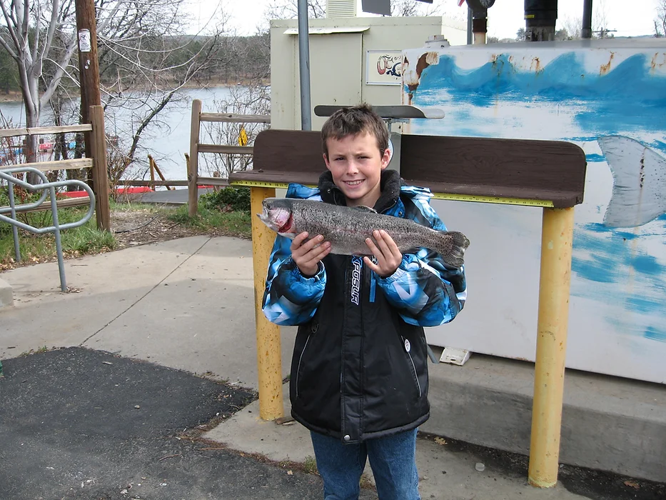 A child in a winter jacket holding a fish outdoors near a lake or river.