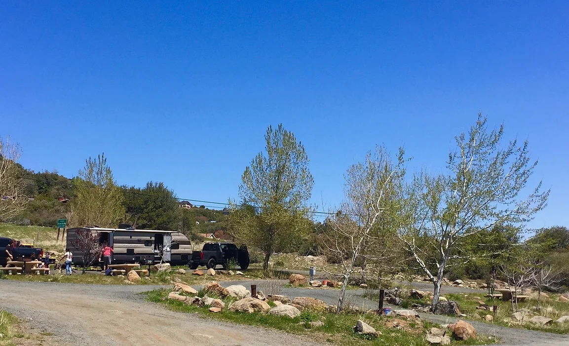 A camper, truck, and people are at a campsite with trees and clear sky in a rural setting.