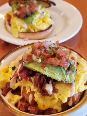 Scrambled eggs, avocado, bacon, and salsa on a bagel and in a bowl, topped with sauce.