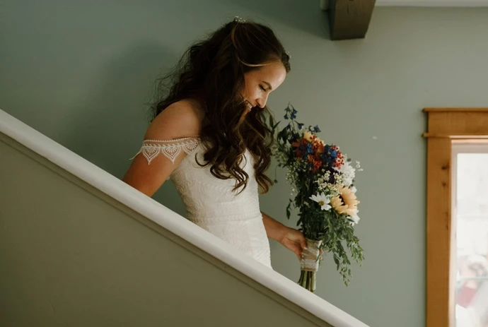 A woman in a white dress descends a staircase holding a bouquet of colorful flowers.