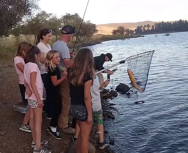A group of people standing by a lake, watching as a man uses a large net to catch a fish near the shore.