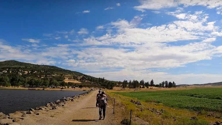 A person walks on a dirt path between a lake and grassy field under a blue sky with clouds.