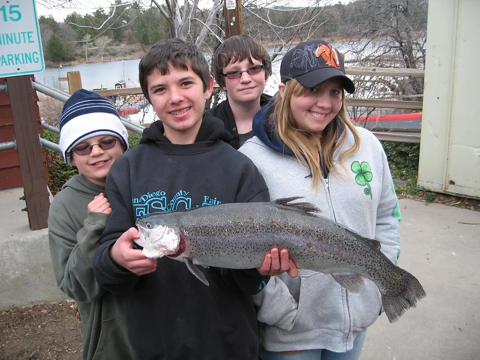 Four people smiling and holding a large fish outdoors near a body of water.