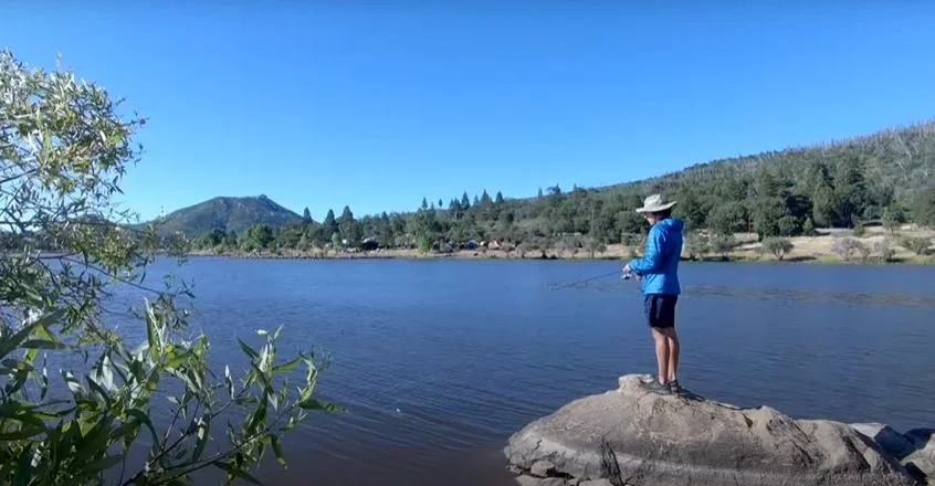A person in a blue jacket and hat is fishing while standing on a rock by a wide lake with a mountainous backdrop.