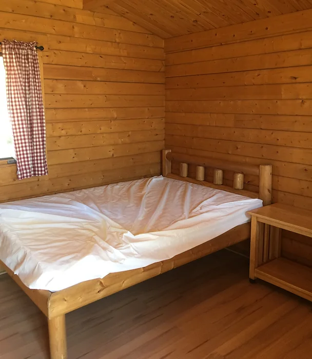 A log cabin room with a wooden bed, white sheets, a small table, and a window with checkered curtains.