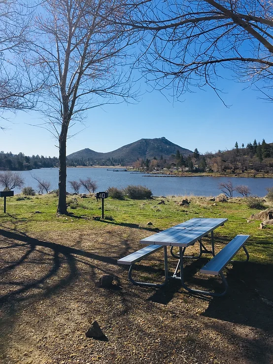 A picnic table near a lake, surrounded by trees and hills under a clear blue sky.