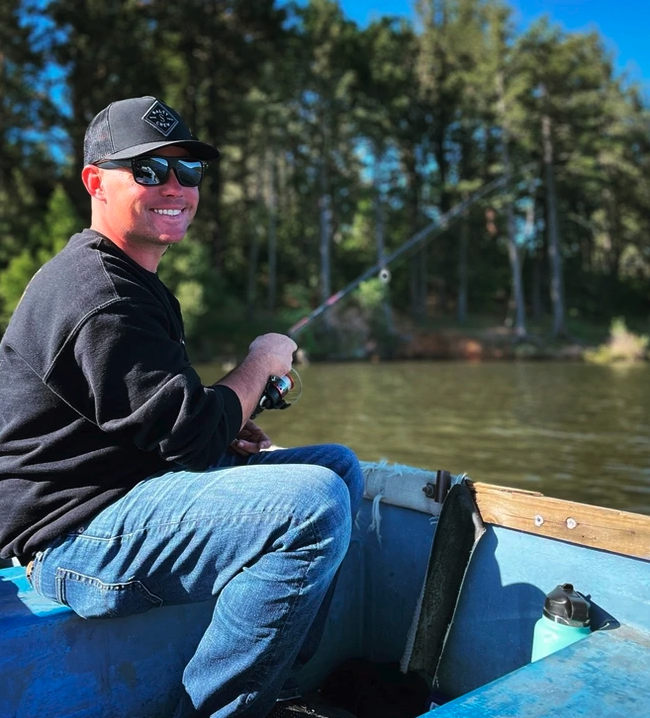 A person in a black cap and sunglasses is fishing on a boat, smiling on a sunny day with trees and water in the background.