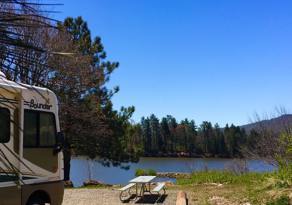 RV parked by a lake with a picnic table, trees, and clear blue sky.