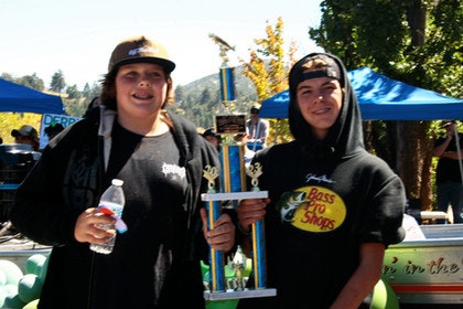 Two people holding a trophy, one with a water bottle, standing outdoors with tents and trees in the background.