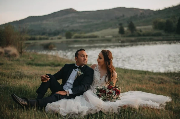 A couple in wedding attire sits on grass by a lake, mountains in the background.
