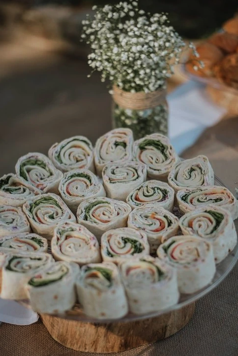 Tortilla rolls with vegetables on a plate, and a vase of small white flowers in the background.