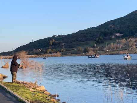 A person fishing by a calm lake with boats and hills in the background.