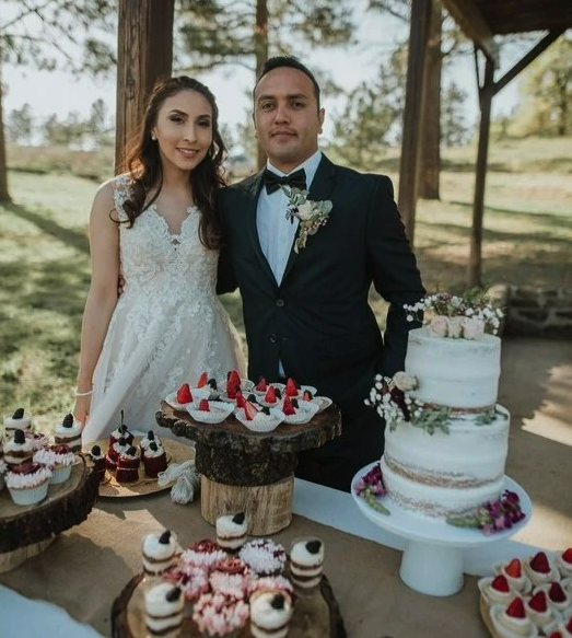 A couple in wedding attire standing by a table of desserts and cake outdoors.