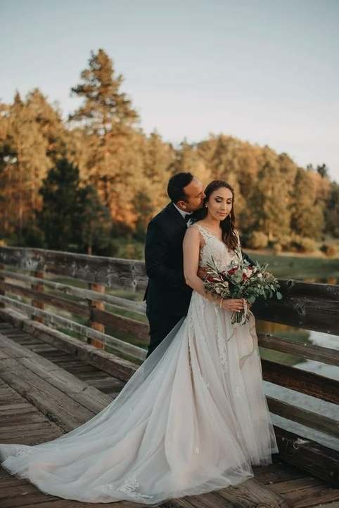 A couple in wedding attire on a wooden bridge, with the groom embracing the bride who is holding a bouquet.