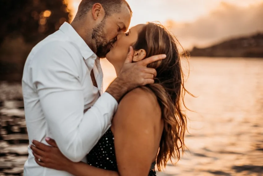 A couple kissing by a lake at sunset, with warm lighting.