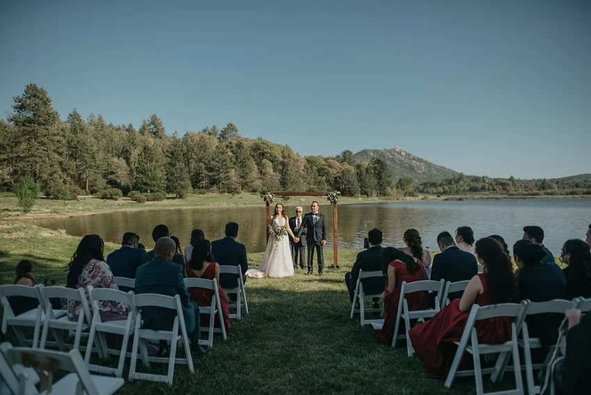 Outdoor wedding ceremony by a lake, with a couple under an arch, surrounded by guests.