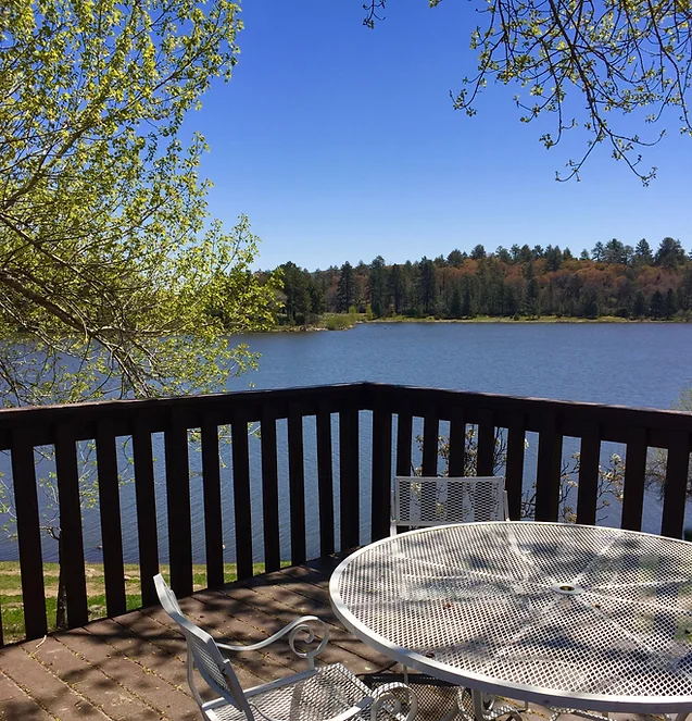 A serene lakeside view with a metal patio table and chairs on a wooden deck, surrounded by trees under a clear blue sky.