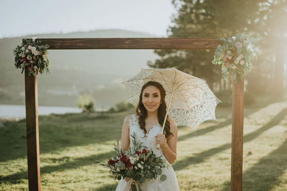 A woman in a white dress holds a parasol and bouquet, standing outdoors under a floral arch.