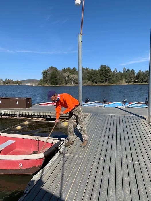 Person in orange shirt pulling a rowboat with a rope at a dock by a lake.