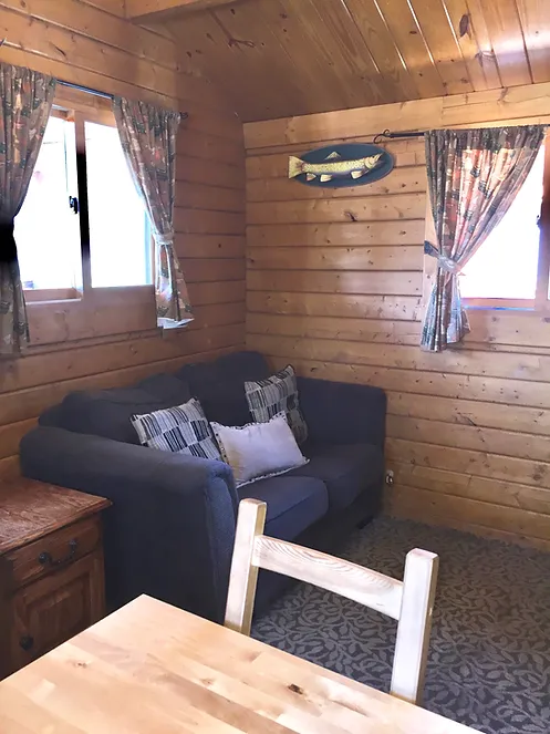 Cozy wooden cabin with a sofa, pillows, curtains, and fish wall decor; natural light from two windows.