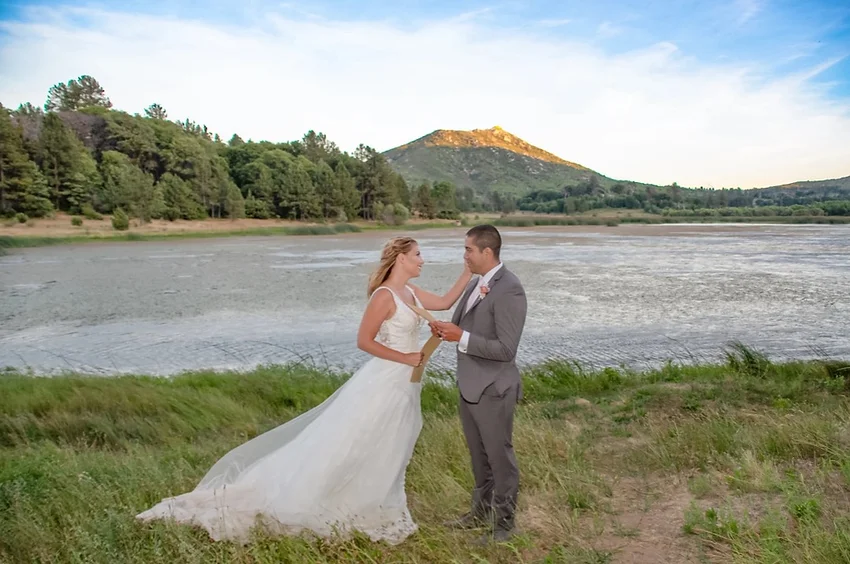 A bride and groom stand by a lake with a mountain backdrop, holding hands.