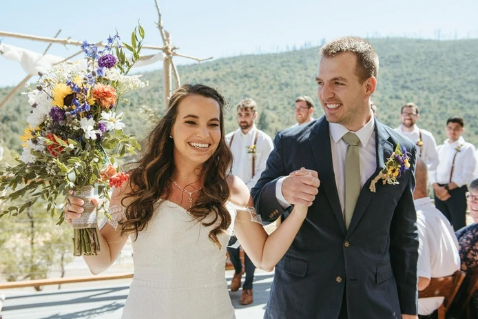 A bride and groom joyfully hold hands, surrounded by their wedding party, with a scenic outdoor backdrop.