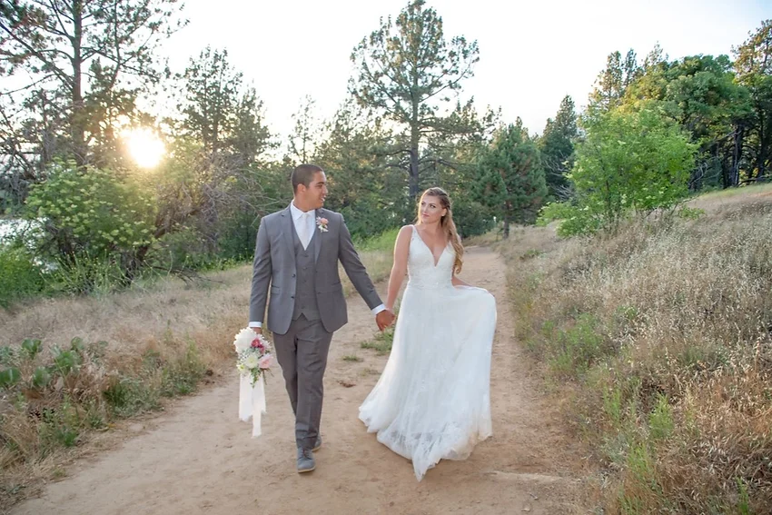 A bride and groom walk hand in hand on a wooded path at sunset, wearing a wedding dress and suit.
