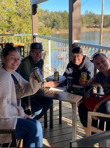 Four people sitting at an outdoor table with drinks, smiling, near a scenic lake.