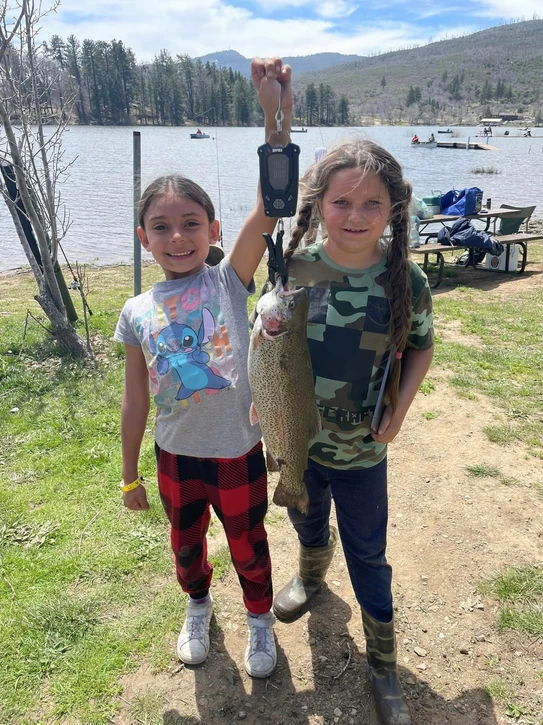 Two kids by a lake, one holding a large fish on a scale.
