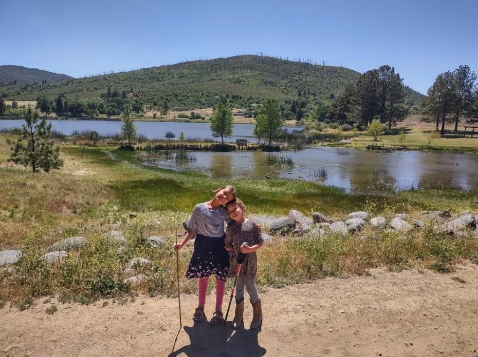 Two children smiling by a pond, with sticks in their hands. There's a grassy area, trees, and hills in the background under a clear sky.