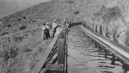 Black and white image of people standing by an aqueduct in a hilly landscape.