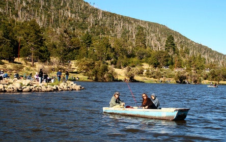 Three people are in a boat fishing on a lake surrounded by trees and hills, with more people on the shore.